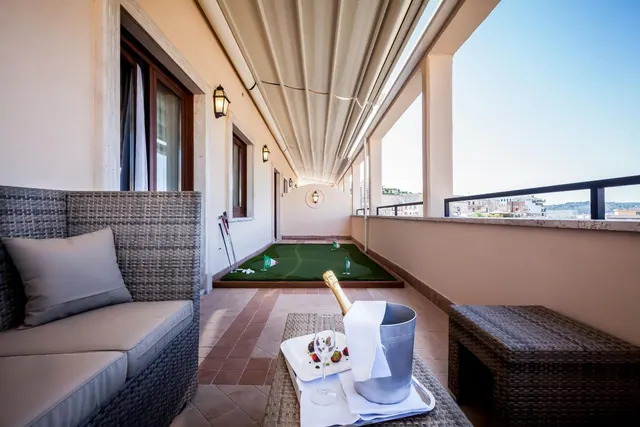 A covered terrace with a wicker sofa, a small putting green, and a table holding a champagne bottle in an ice bucket and a plate of fruit, with rooftops visible beyond the railing.