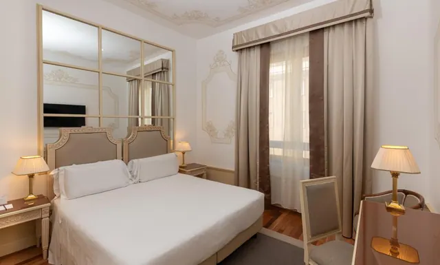 A bedroom with a double bed, a mirrored wall panel behind the headboard, bedside lamps, a curtained window, an upholstered chair and a console table with a lamp.
