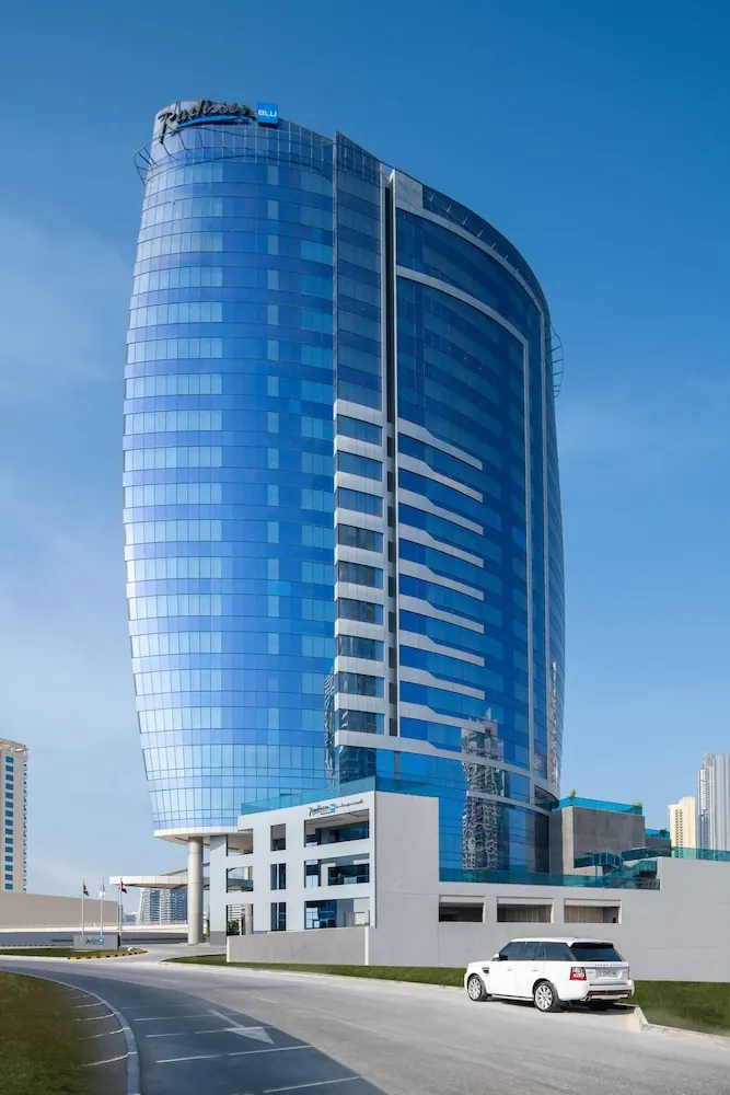 A curved blue glass hotel tower with a Radisson Blu sign on its roof rises under a clear sky, with a white SUV parked on the driveway below.
