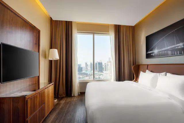 A bedroom with a large white bed, a wall-mounted television, and a window looking out over a city skyline and waterway.