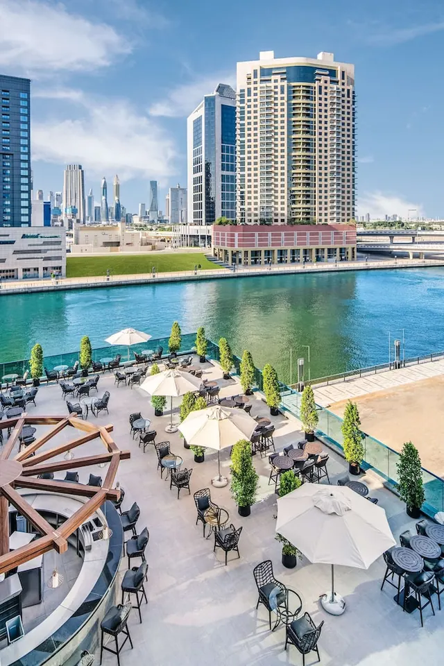 An outdoor terrace with tables, chairs and white umbrellas overlooks a canal with high-rise towers across the water.