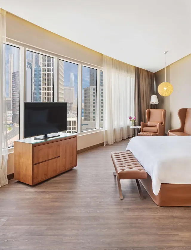 A hotel bedroom with a king bed, a tufted bench, two armchairs, a wooden TV console and floor-to-ceiling windows overlooking high-rise buildings.