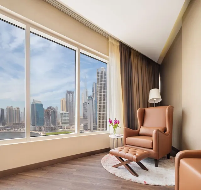 A seating area with a tan leather armchair, ottoman and floor lamp beside large windows looking out over a city skyline with high-rise towers.