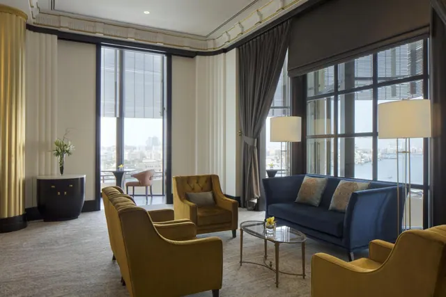 A suite living area with gold armchairs, a navy sofa, a glass coffee table and tall windows looking out over water and city buildings.