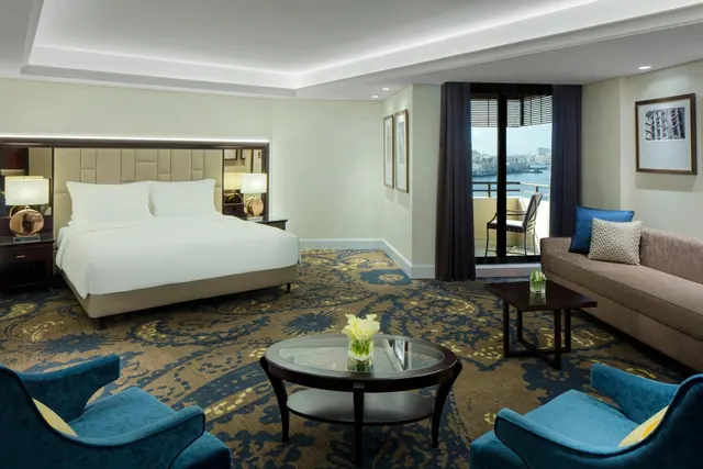 A hotel suite bedroom with a king bed, a seating area with a sofa and armchairs, and a glass door opening onto a balcony with a view of water and buildings.