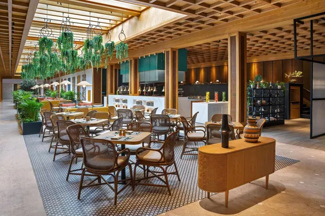 A restaurant dining room with rattan chairs and wooden tables under a lattice wood ceiling, with hanging plants overhead and a kitchen counter in the background.