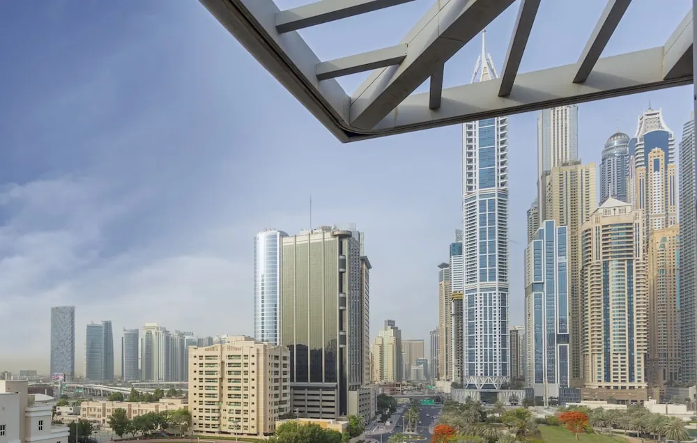 View past a metal overhead frame across a dense cluster of high-rise towers and a tree-lined road below.