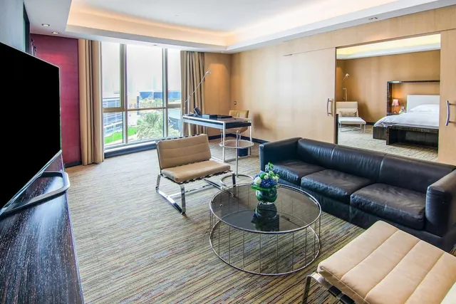A furnished living area in a hotel suite with a black leather sofa, armchairs, a round glass coffee table, a desk, a wall-mounted television, and a view of the bedroom through an open sliding door.
