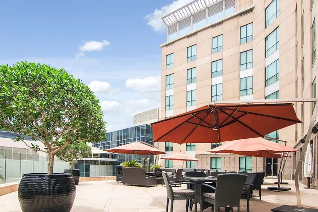 An outdoor terrace with red parasol umbrellas, wicker dining sets, large planters with trees, and a multi-storey hotel building beside it.