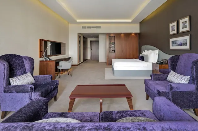 A hotel suite living area with purple velvet armchairs and a sofa around a low wooden coffee table, with a double bed visible in the background.