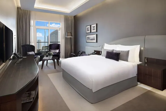 A king bed with white linens and purple cushions sits in a bedroom with an armchair seating area, a wall-mounted television, and a window view of a tall spired tower and city buildings.