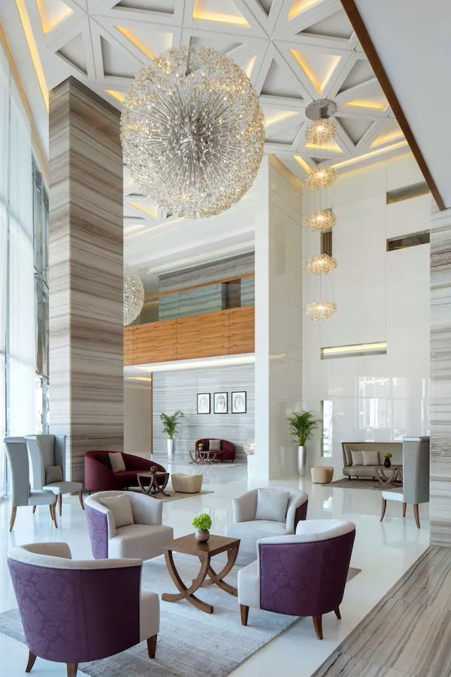 A lobby with a large crystal chandelier, purple armchairs grouped around a wooden coffee table, and marble columns beneath a geometric ceiling.