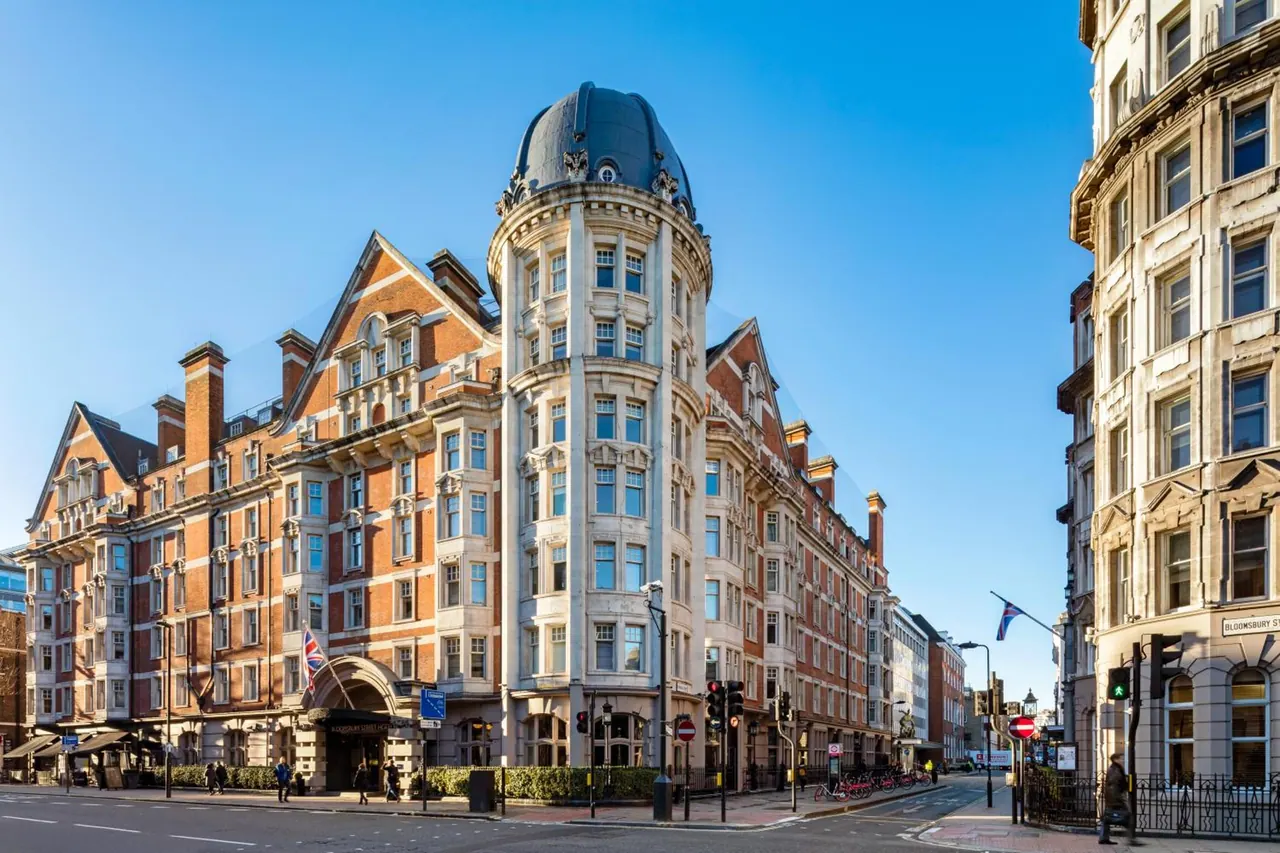Exterior of a red-brick Victorian hotel with a domed corner tower and a Union Jack flag, beside a street with pedestrians, traffic lights and parked bicycles under a clear blue sky.