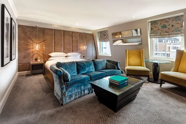 A hotel bedroom with a double bed, a blue velvet sofa, a glass-topped coffee table, two yellow armchairs and a window with a floral roman blind looking onto neighbouring buildings.