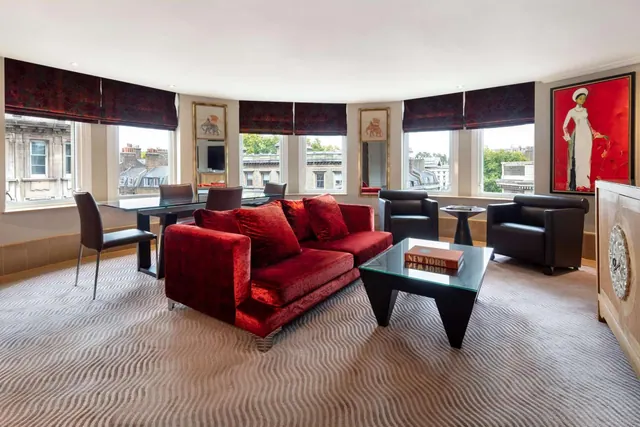 A living room with a red velvet sofa, black leather armchairs, a glass coffee table and a glass-topped dining table in front of curved windows overlooking rooftops.
