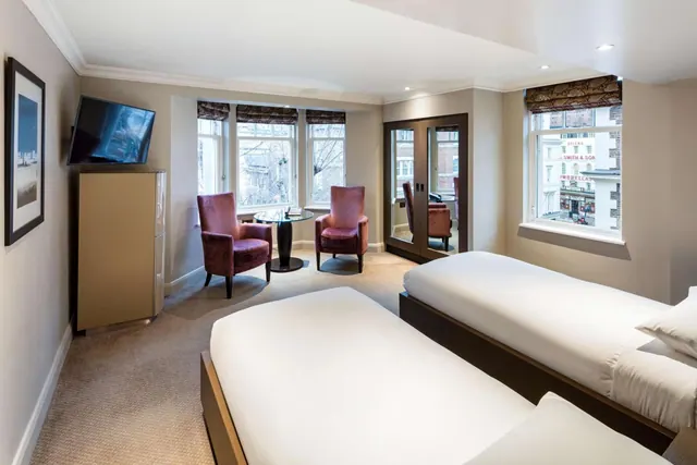 A bedroom with two single beds, two red armchairs beside a round glass table, a wall-mounted television, and a bay window looking onto a street with buildings.