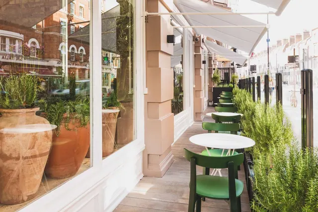 An outdoor terrace with green chairs, round white tables, canvas awnings and potted rosemary along the edge, with a street and terraced houses visible beyond the railings.