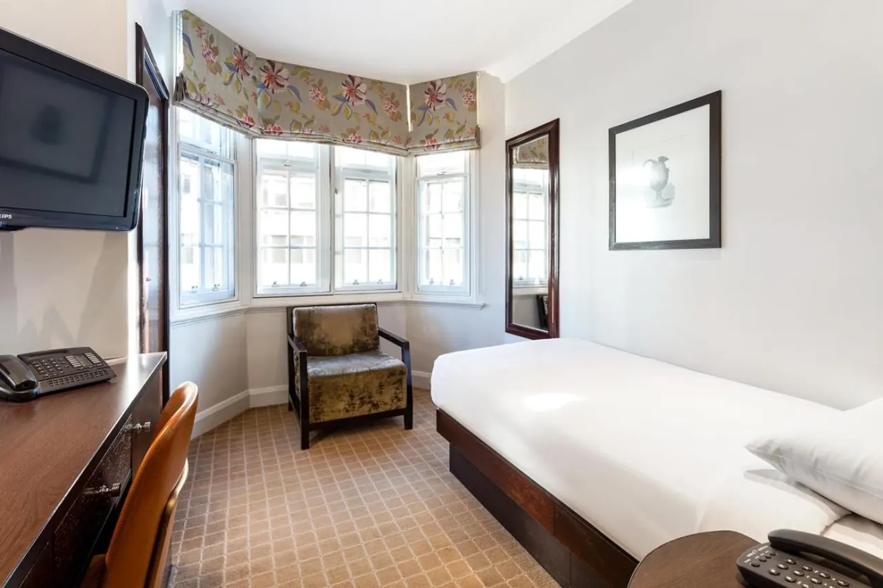 A single bed sits against a white wall beside a bay window with a floral roman blind, an armchair, a wall-mounted television and a desk with a telephone.