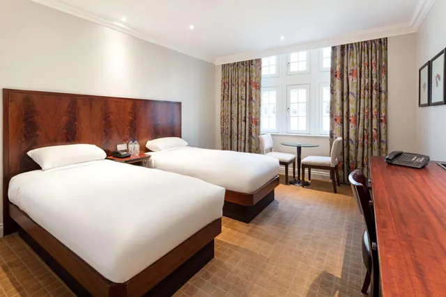 A twin bedroom with two single beds with wooden headboards, a small round table with two chairs by a window with floral curtains, and a wooden desk with a telephone on the right.