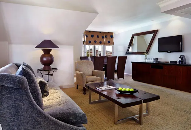 A living area with a grey sofa, armchair, wooden coffee table holding a bowl of green apples, wall-mounted television, wooden sideboard with a coffee machine, and a dining table by the windows.