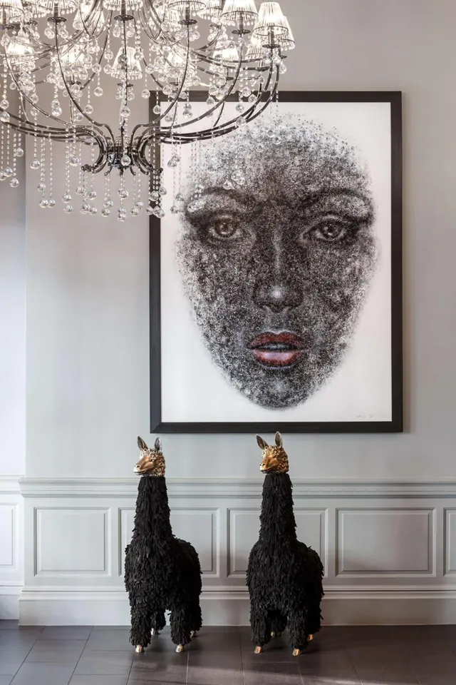 A hallway or lobby with a crystal chandelier, a large framed portrait of a face made of dots, and two black llama sculptures with gold heads on a dark tiled floor.