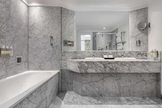 A marble-tiled bathroom with a built-in bathtub, a double vanity with two sinks, and a glass-enclosed shower visible in the mirror.