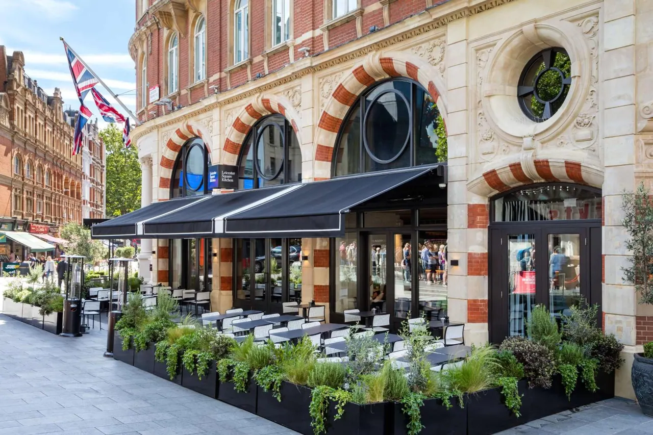 The exterior of a brick and stone building with striped arches and a black awning, with an outdoor dining area of tables and planters on the pavement.