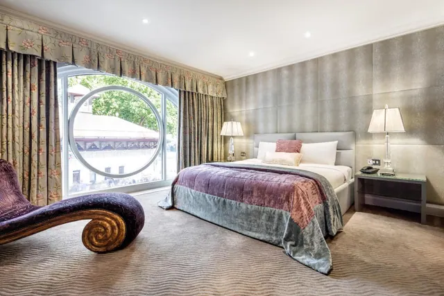A bedroom with a double bed, a chaise longue, two bedside lamps and a large round-framed window with floral curtains.
