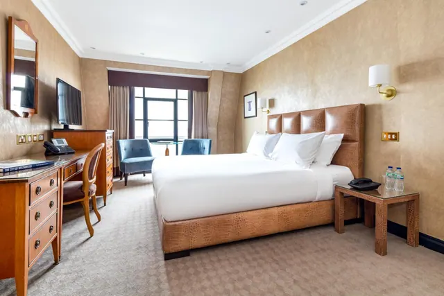 A hotel bedroom with a large bed with a brown leather headboard, a wooden desk and dresser, blue armchairs by a window, and a wall-mounted television.