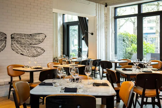 A restaurant dining room with white brick walls, a dragonfly wing mural, marble-topped tables set with wine glasses and tan leather chairs, and large windows looking out onto trees.