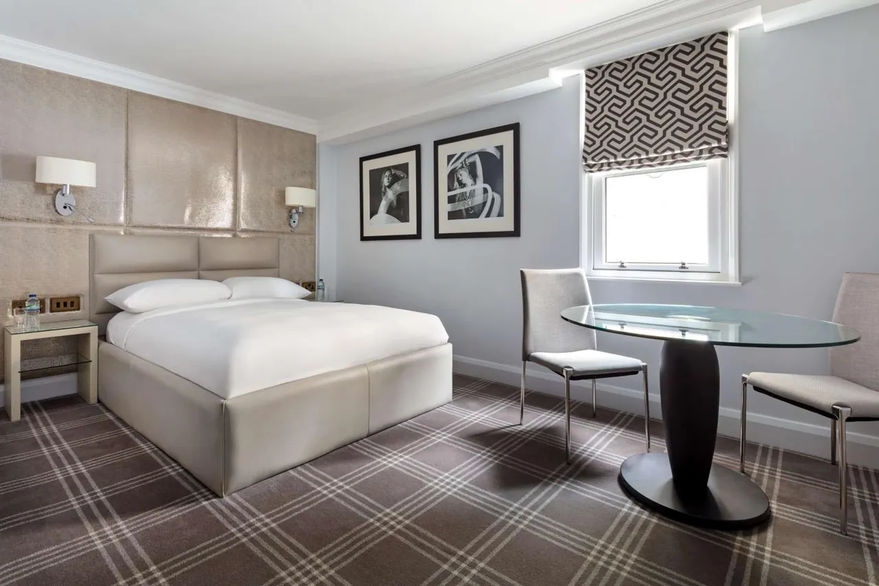 A hotel bedroom with a made-up double bed, a round glass table with two chairs, and a window with a patterned roman blind.