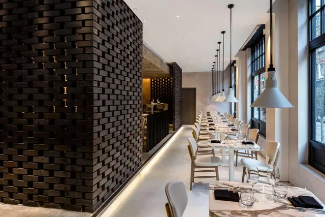 A long restaurant dining room with white upholstered chairs and marble-topped tables set with dark napkins and glasses, beside a dark brick feature wall and under white pendant lamps.