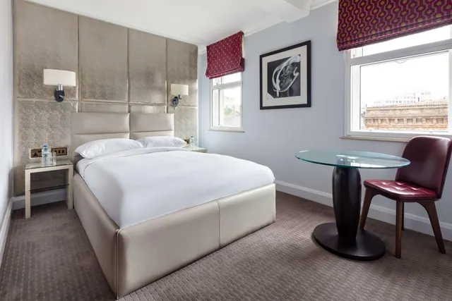 A made-up double bed with a padded headboard sits beside a round glass table and chair, with windows looking out onto buildings.