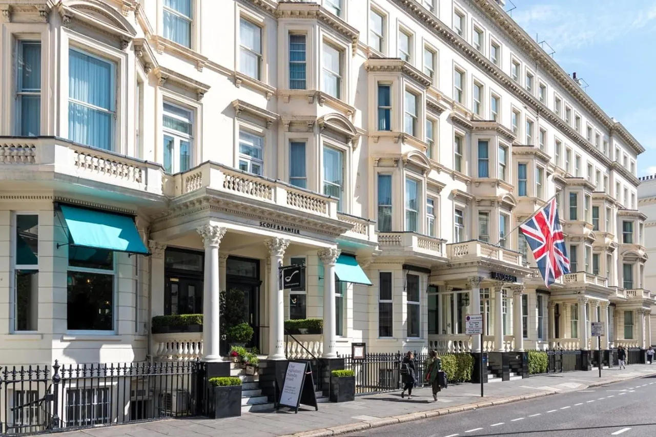 A cream stucco facade of a hotel on a London street, with teal awnings, a Union Jack flag and pedestrians on the pavement.