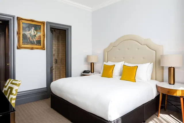 A bedroom with a made king bed, yellow cushions, a framed painting on the wall, and a glass-walled shower visible through an open doorway.