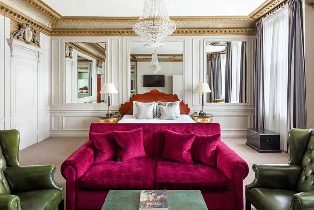 A suite with a red velvet sofa, green leather armchairs, a wooden-headboarded bed and a crystal chandelier.