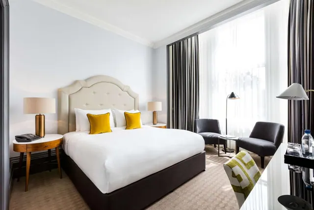 A bedroom with a king bed with yellow cushions, two armchairs by a tall window with sheer curtains, and a desk on the right.