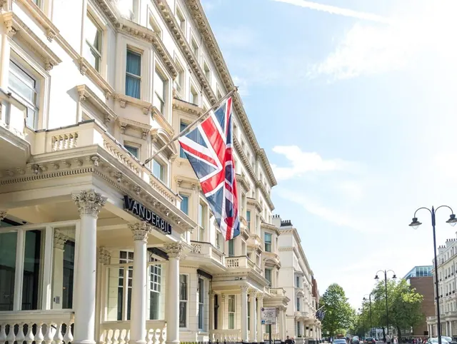 Cream stucco facade of a building with a Vanderbilt sign and a Union Jack flag, beside a tree-lined street with parked cars.