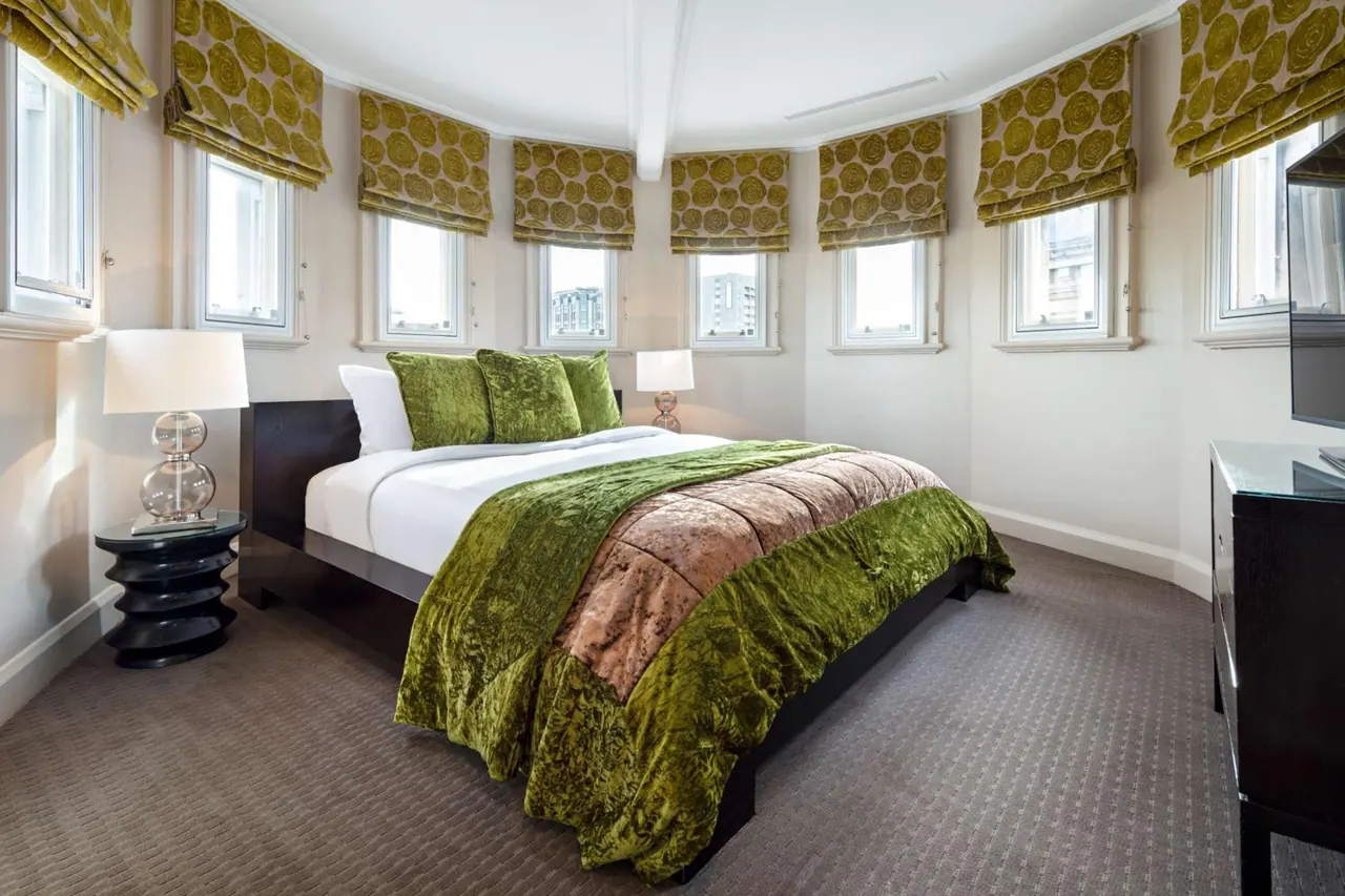 A bedroom with a king bed under green velvet throws, a curved wall of windows with patterned roman blinds, and a dark dresser with a television on the right.