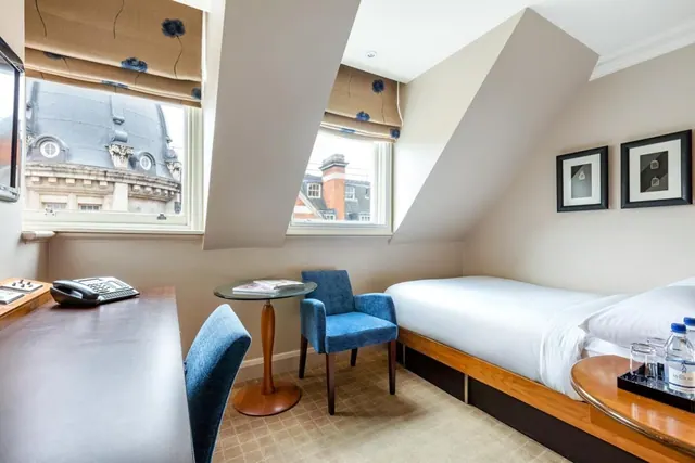 A sloped-ceiling hotel bedroom with a single bed, a desk with a telephone, two blue armchairs and a view of rooftops through two windows.