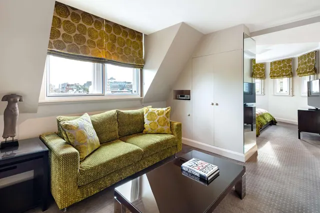 A living area with a green patterned sofa, a window with patterned roman blinds looking out over city rooftops, and a bedroom with a television visible through an opening on the right.