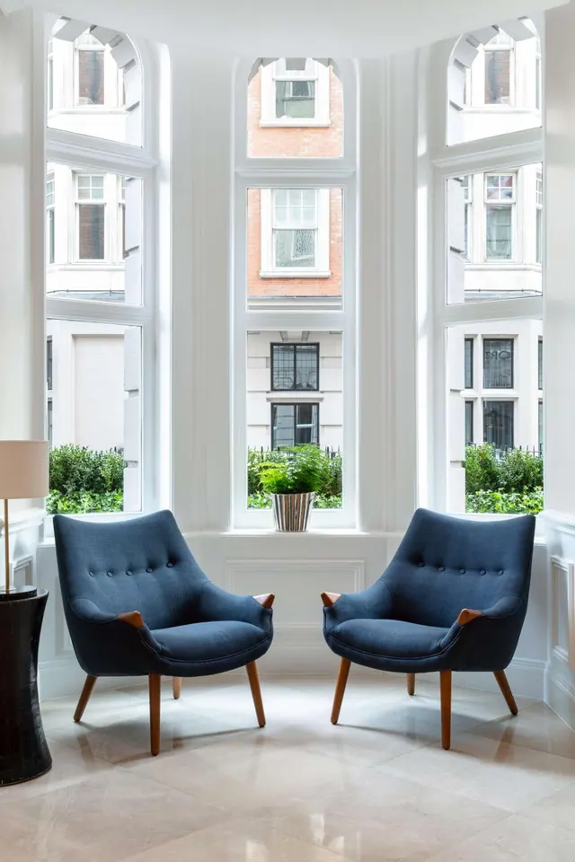Two blue upholstered armchairs stand in a white bay window with a potted plant on the sill, looking out onto neighbouring brick and stone buildings and hedges.