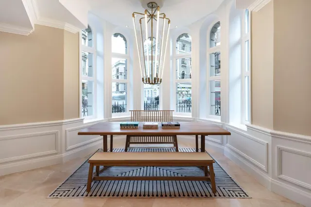 A wooden dining table with a bench and chair sits under a brass chandelier in a bay window nook, with buildings and railings visible outside.