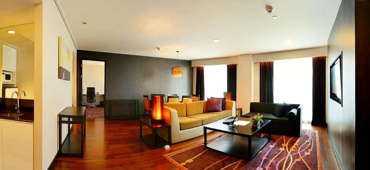 A suite living area with beige and dark sofas around a low coffee table, a kitchenette on the left, a desk visible through a doorway, a wall-mounted television, and tall windows with curtains.