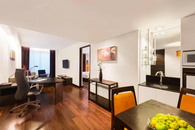 A hotel suite living area with a desk and office chair, a sofa, a wall-mounted television, a kitchenette with a sink and microwave, and a dining table with a fruit bowl, with the bedroom visible through a doorway.