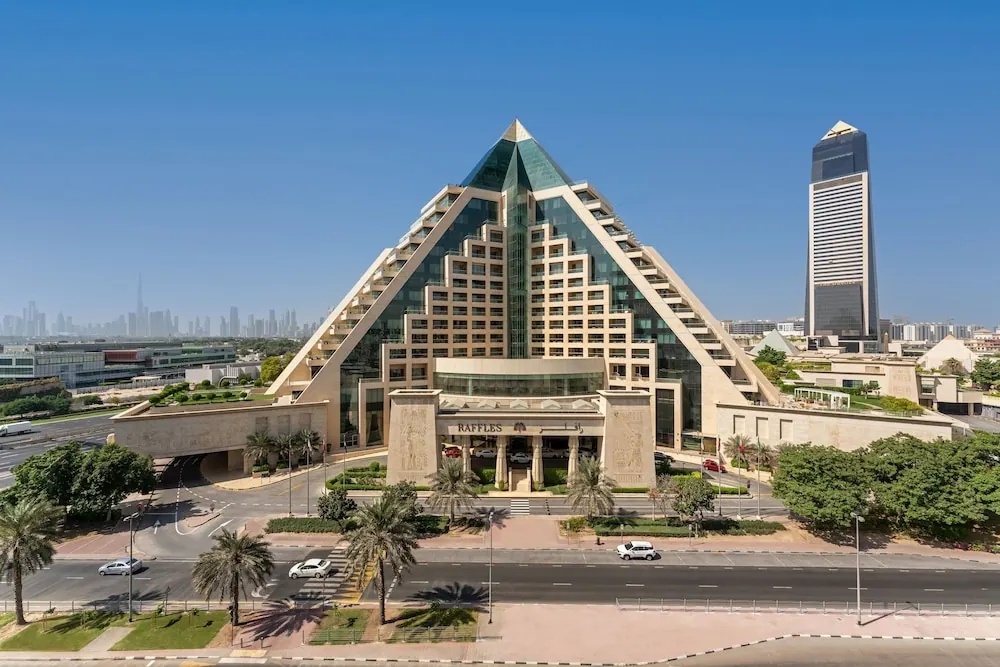 Exterior view of the Raffles Dubai hotel, a pyramid-shaped tower, with a road, palm trees and a tall office tower beside it under a clear blue sky.