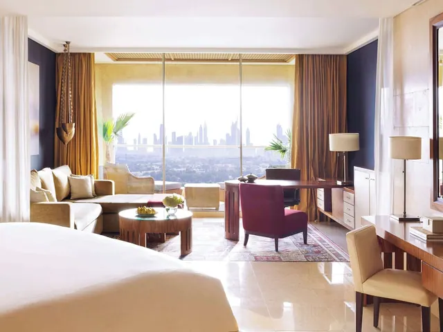 A hotel suite bedroom with a large bed, a seating area and a desk, with floor-to-ceiling windows looking out over a hazy city skyline.
