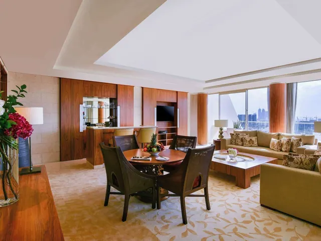A hotel suite living area with a round dining table, sofas, a wall-mounted television and floor-to-ceiling windows overlooking a city skyline.