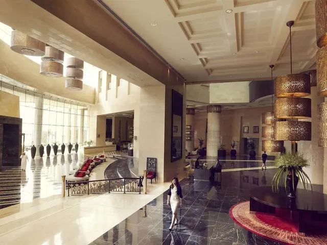 A large hotel lobby with a coffered ceiling, pendant lamps, a curved row of red-cushioned seating, and a woman walking across the marble floor.