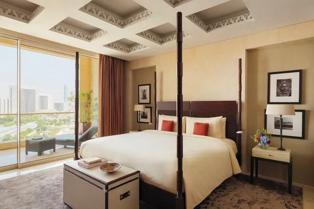 A bedroom with a four-poster king bed and a coffered ceiling, with sliding glass doors opening onto a balcony with city buildings and greenery beyond.